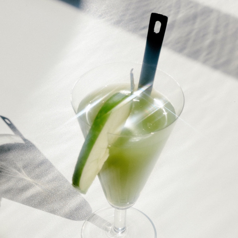 Green cocktail in a tall glass garnished with a cucumber slice and a black stirrer, casting a shadow on a white surface.