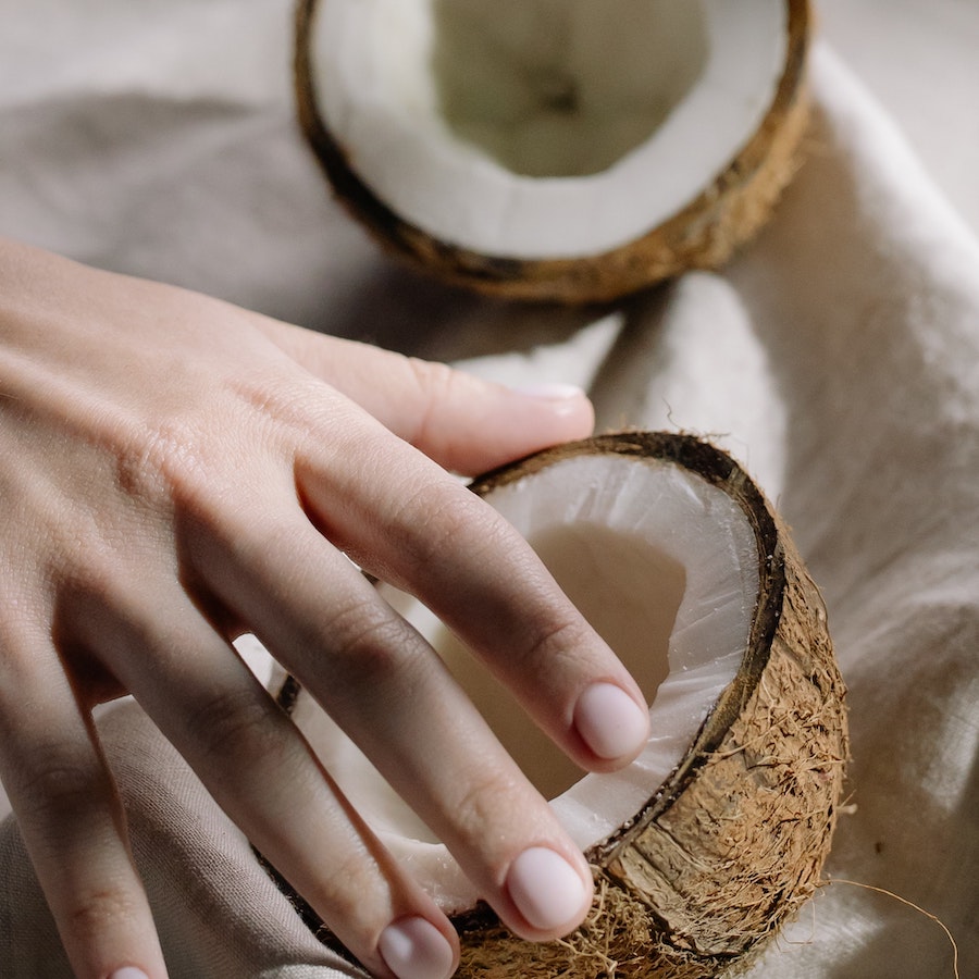 Hand holding a halved coconut with white flesh visible, resting on a beige cloth with another halved coconut in the background.