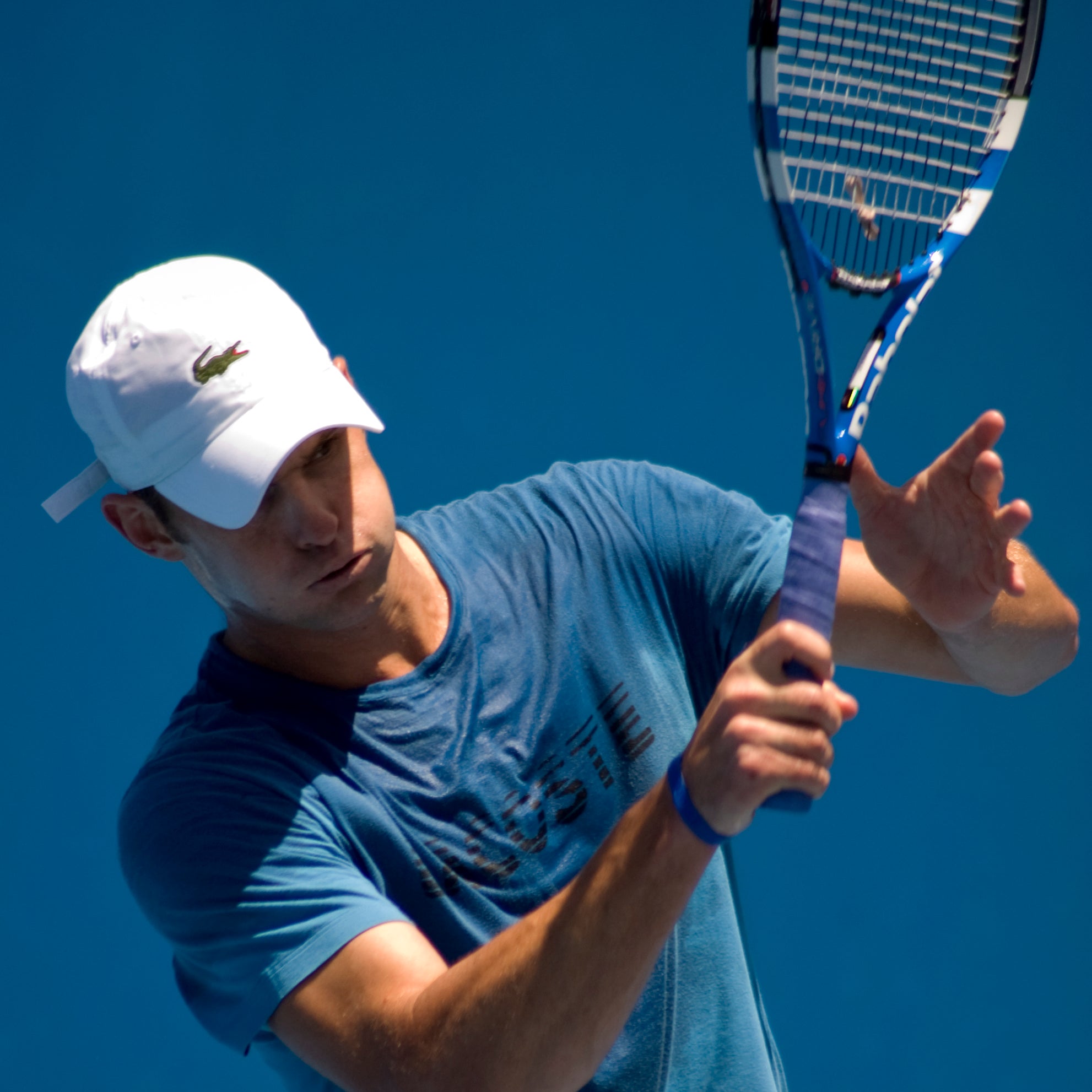 Tennis player swinging a racket during practice, wearing a white cap and blue shirt against a clear blue sky