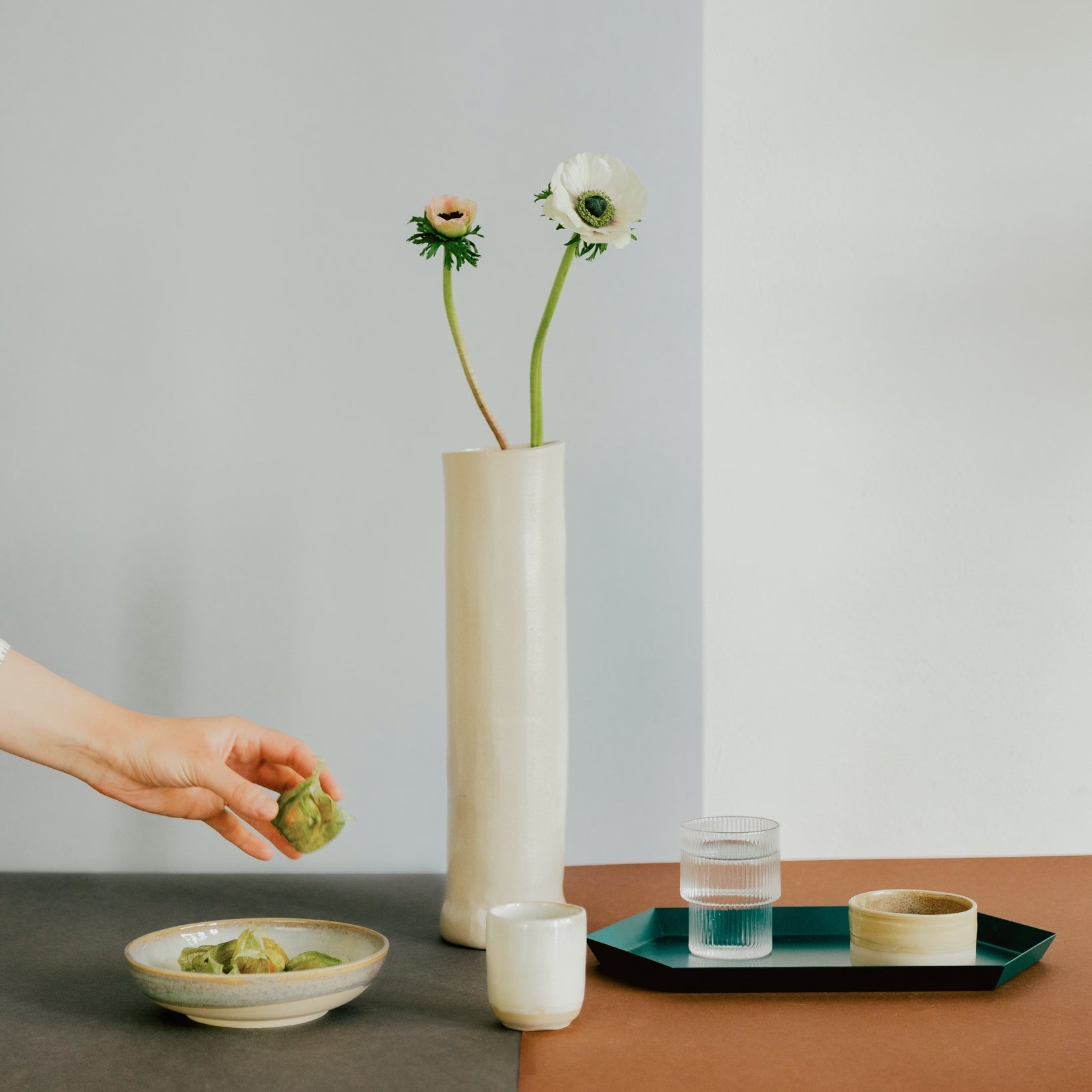 Hand holding a green husk above a bowl with more husks, next to a tall beige vase with two flowers, a white cup, and a glass of water on a teal tray on a brown and gray surface.