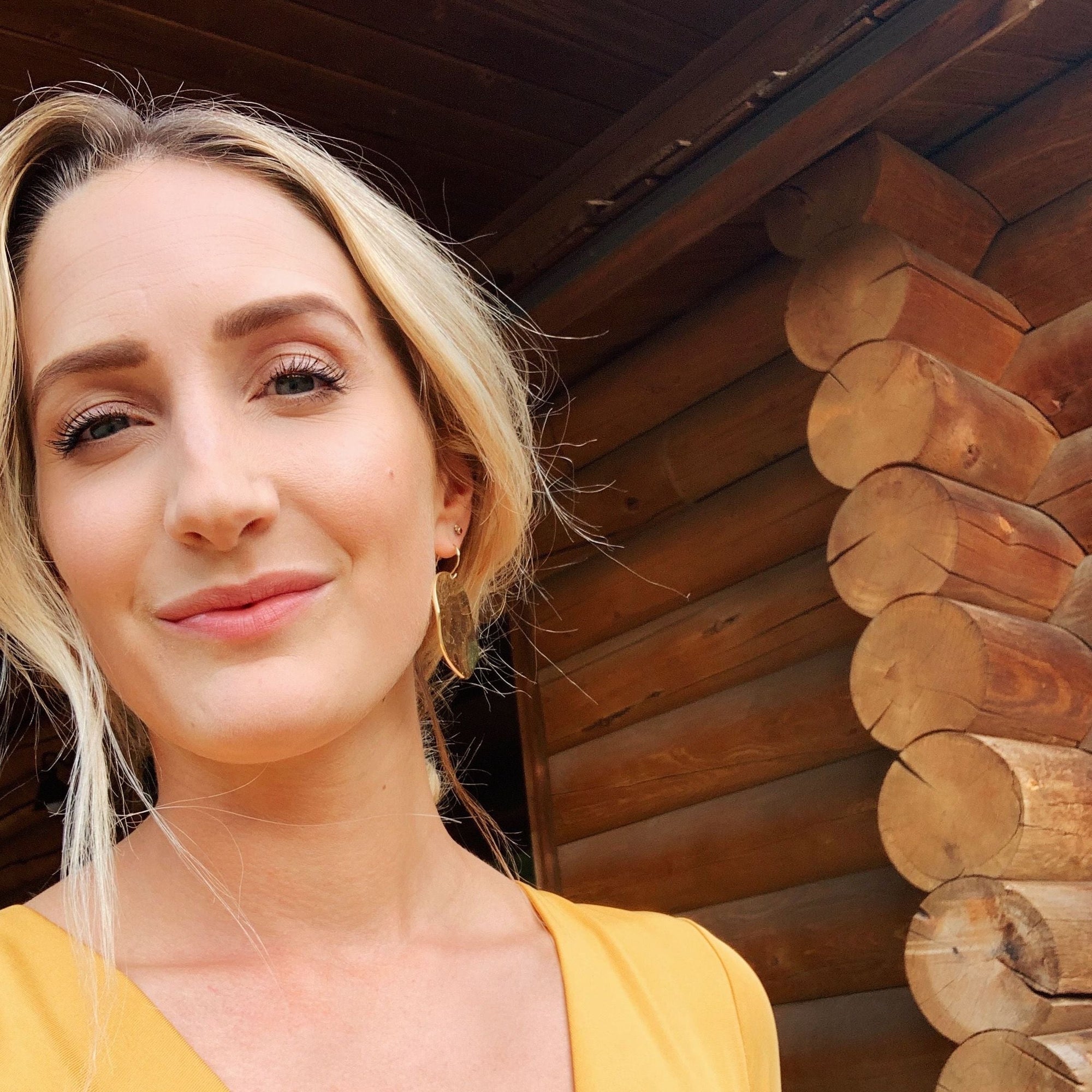 Close-up of a smiling woman in a yellow top in front of a log cabin exterior