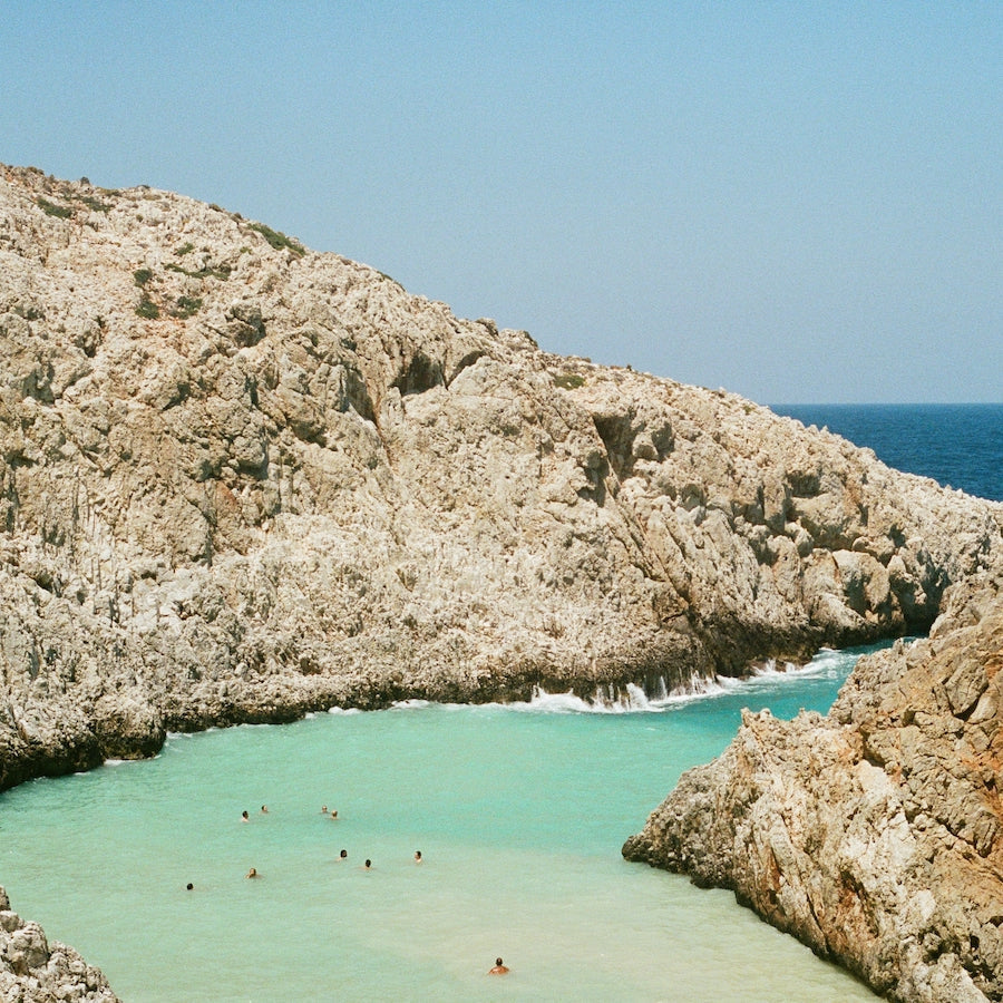Rocky coastal cove with turquoise water and several people swimming near the shore under a clear blue sky