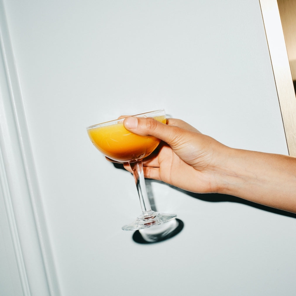 Hand holding a coupe glass filled with an orange mocktail against a white wall