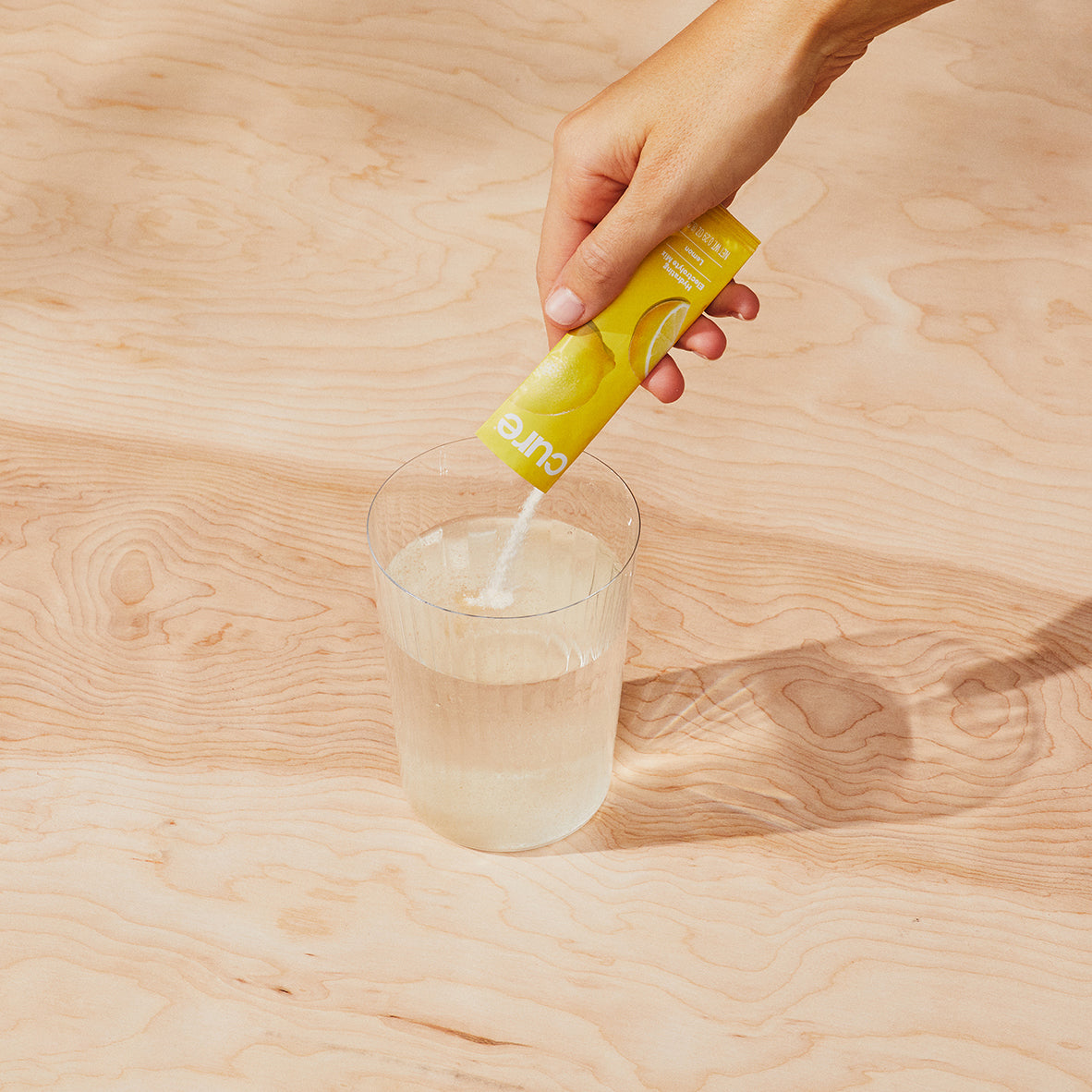 Hand pouring lemon-flavored Cure hydration electrolyte mix into a clear glass of water on a wooden surface