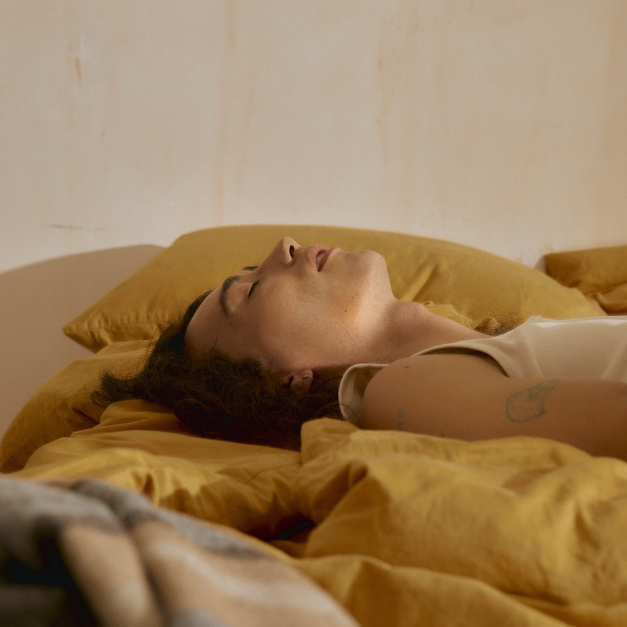 Adult lying on their back with eyes closed on mustard-yellow bedding, appearing fatigued and unwell