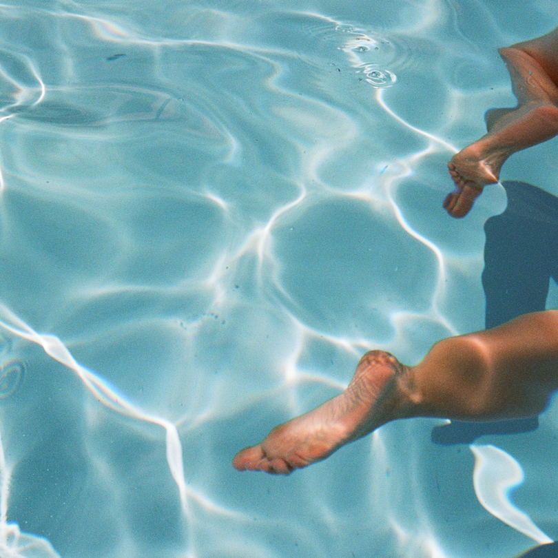 Person's legs and feet floating in a sunlit pool with rippling water reflections