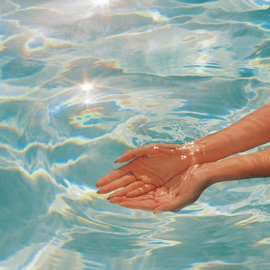 Two hands cupped under clear water with sunlight creating sparkling reflections on the skin and water surface.