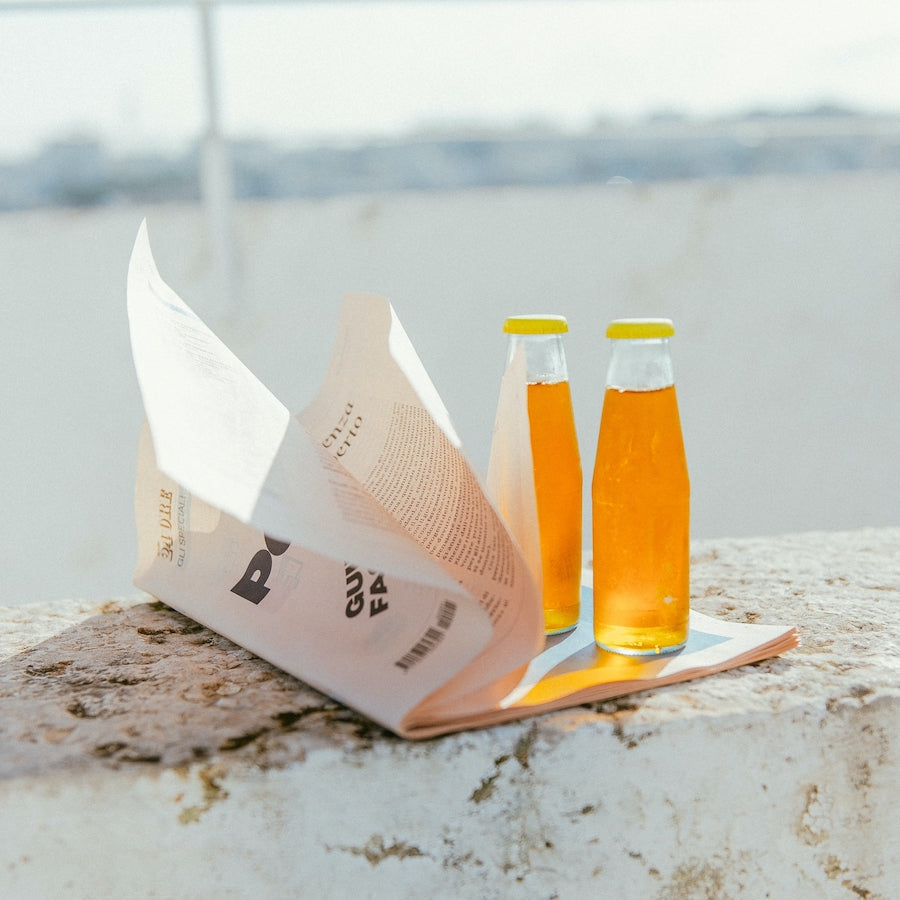 Two glass bottles with orange liquid and a partially folded newspaper on a stone surface in bright daylight.