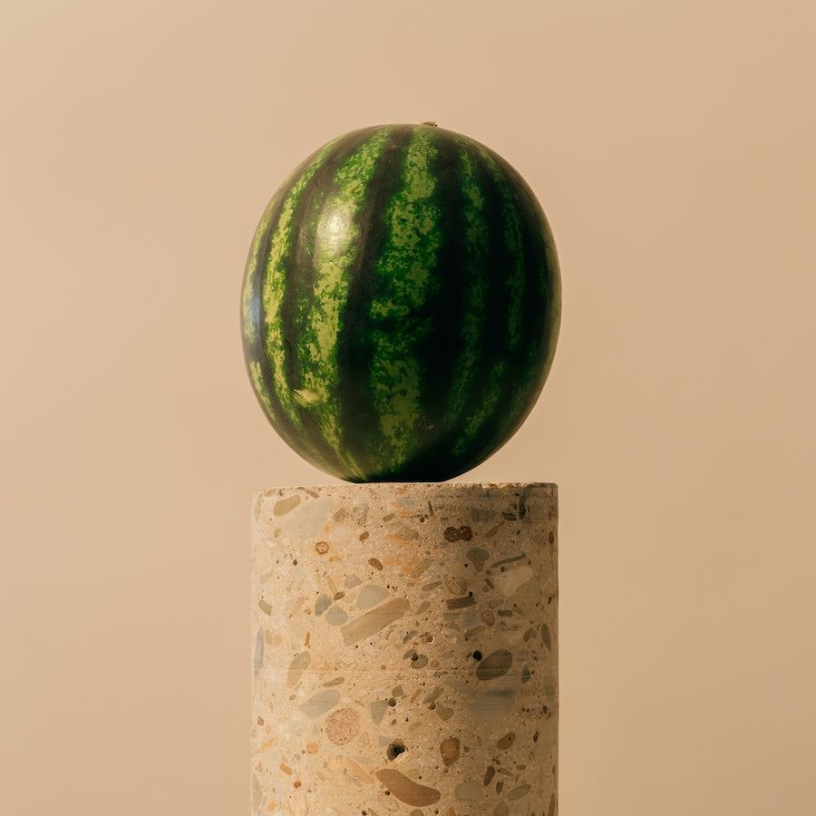 Watermelon with dark and light green stripes balanced on a cylindrical stone pedestal against a beige background