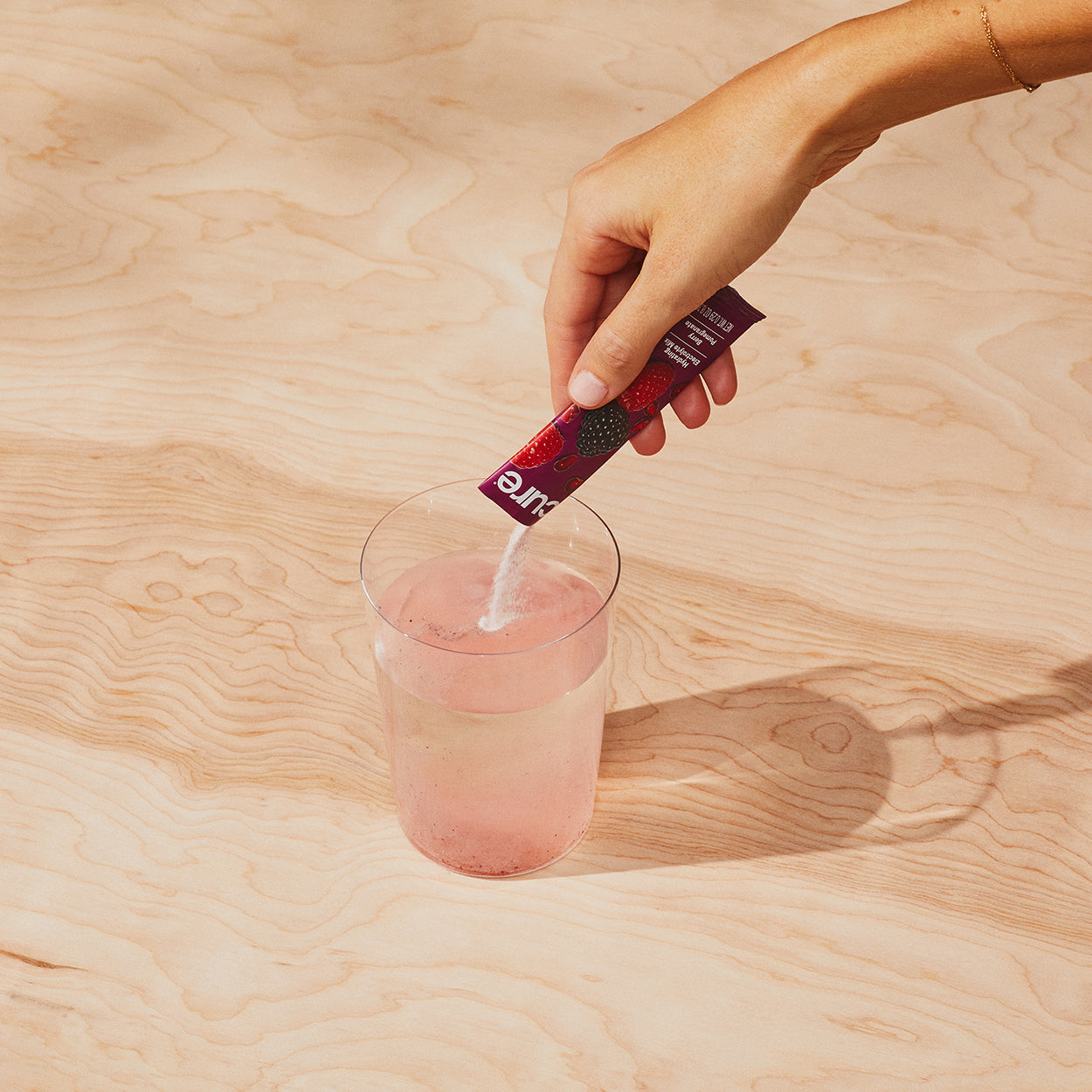 Hand pouring powder from a Cure packet labeled 'Cure' with images of berries into a glass of water on a wooden surface