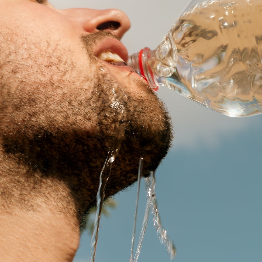 Man drinking water from a plastic bottle with water spilling down his chin against a blue sky background