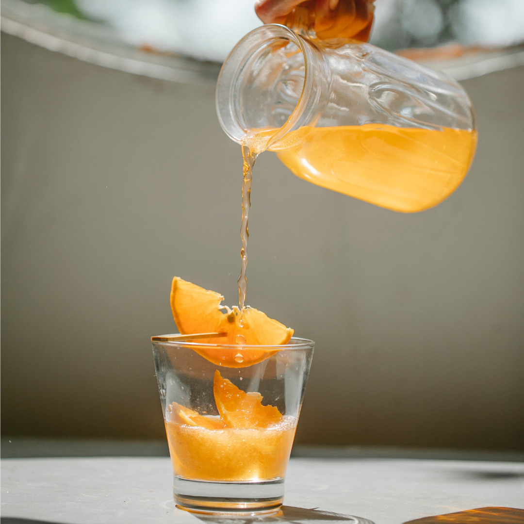 Orange juice being poured from a glass pitcher into a tumbler with orange wedges in soft natural light
