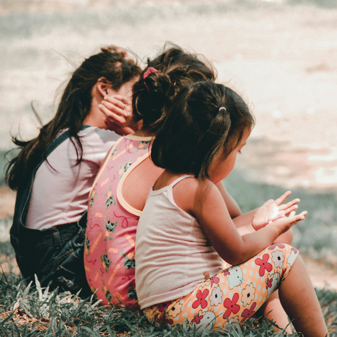 Three young children sitting on grass outdoors, viewed from behind, wearing colorful clothing.