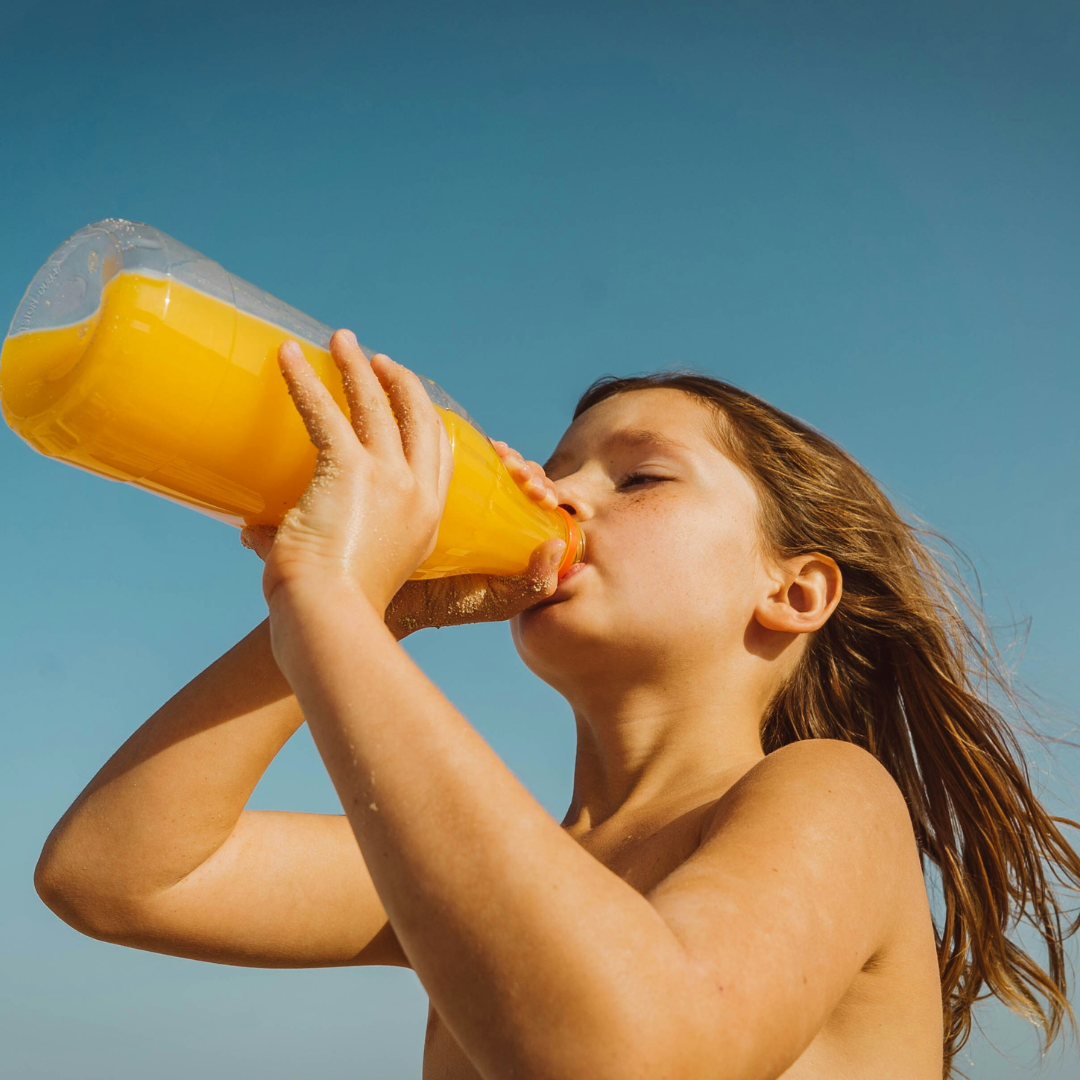 Child drinking orange juice from a clear bottle outdoors against a blue sky
