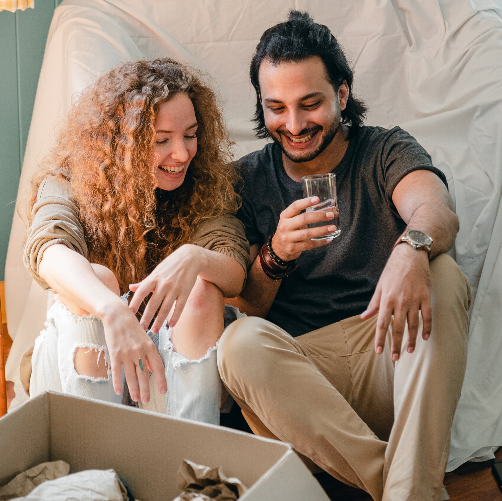 Smiling young couple sitting on the floor unpacking a box; man holding a glass of water, woman with curly hair in ripped jeans.