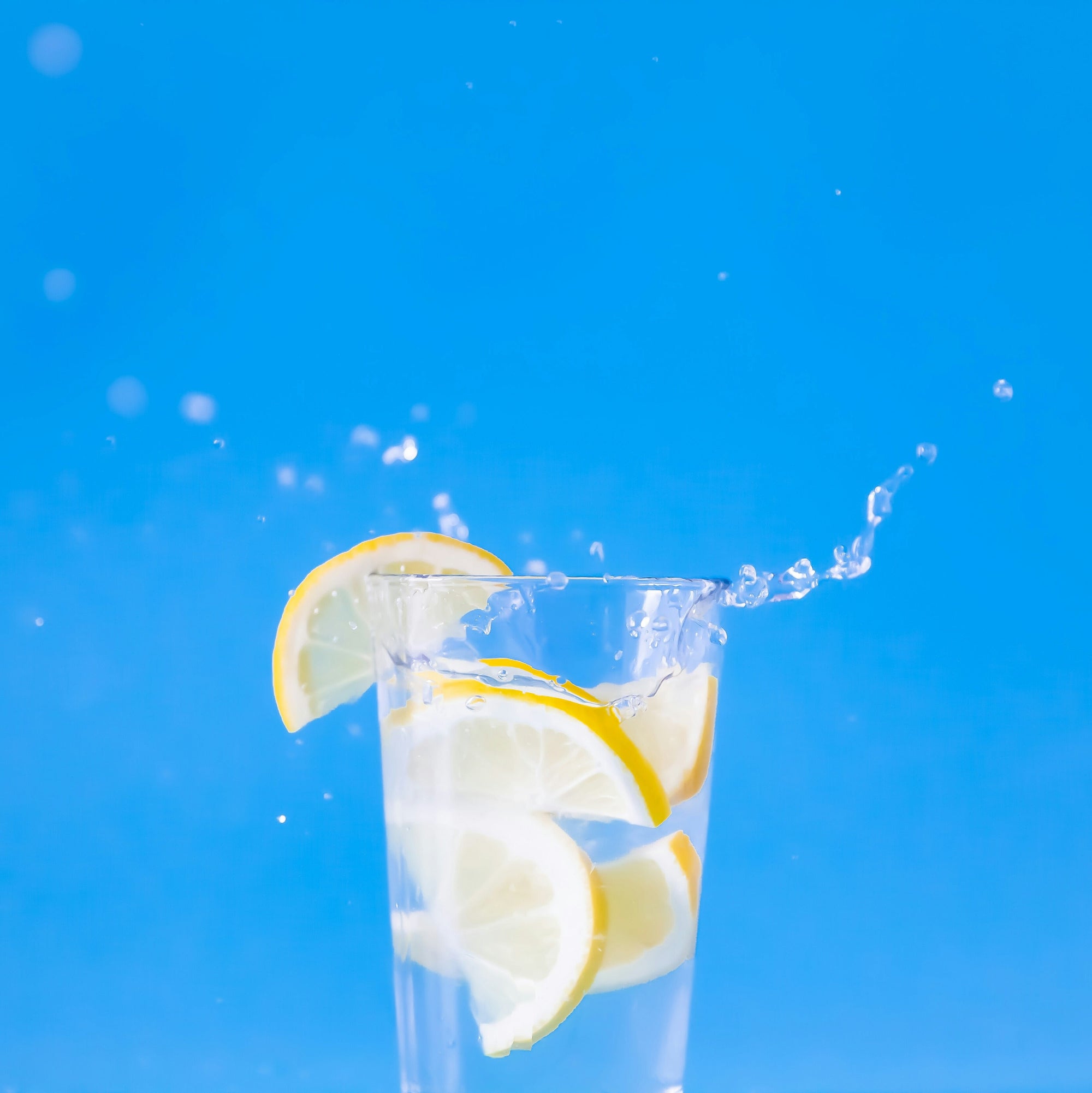 Glass of water with lemon slices splashing against a bright blue background.