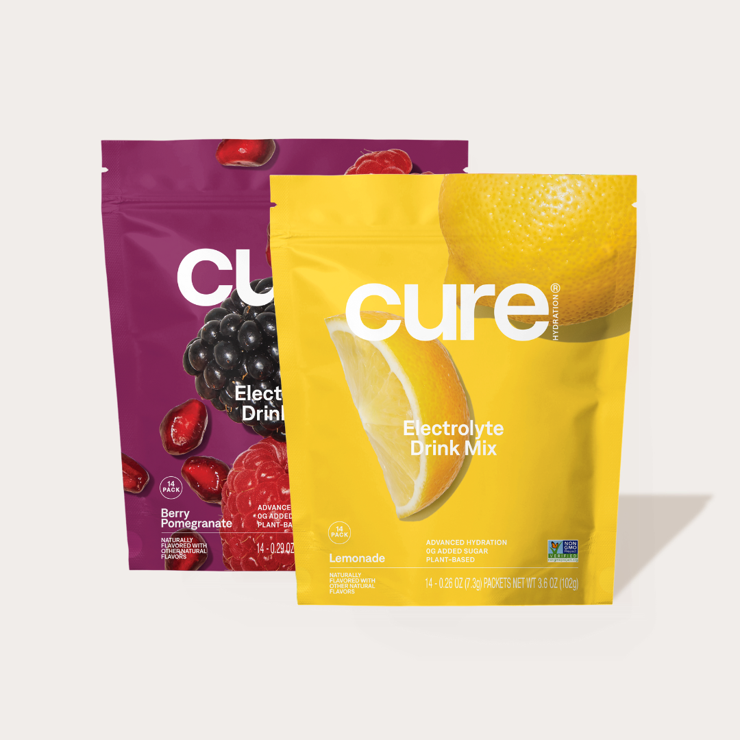 Two Cure Electrolyte Drink Mix packets: one Berry Pomegranate flavor with images of pomegranate seeds and blackberries, and one Lemonade flavor with a lemon slice image. Both show 'Electrolyte Drink Mix' and 'All Natural' text.