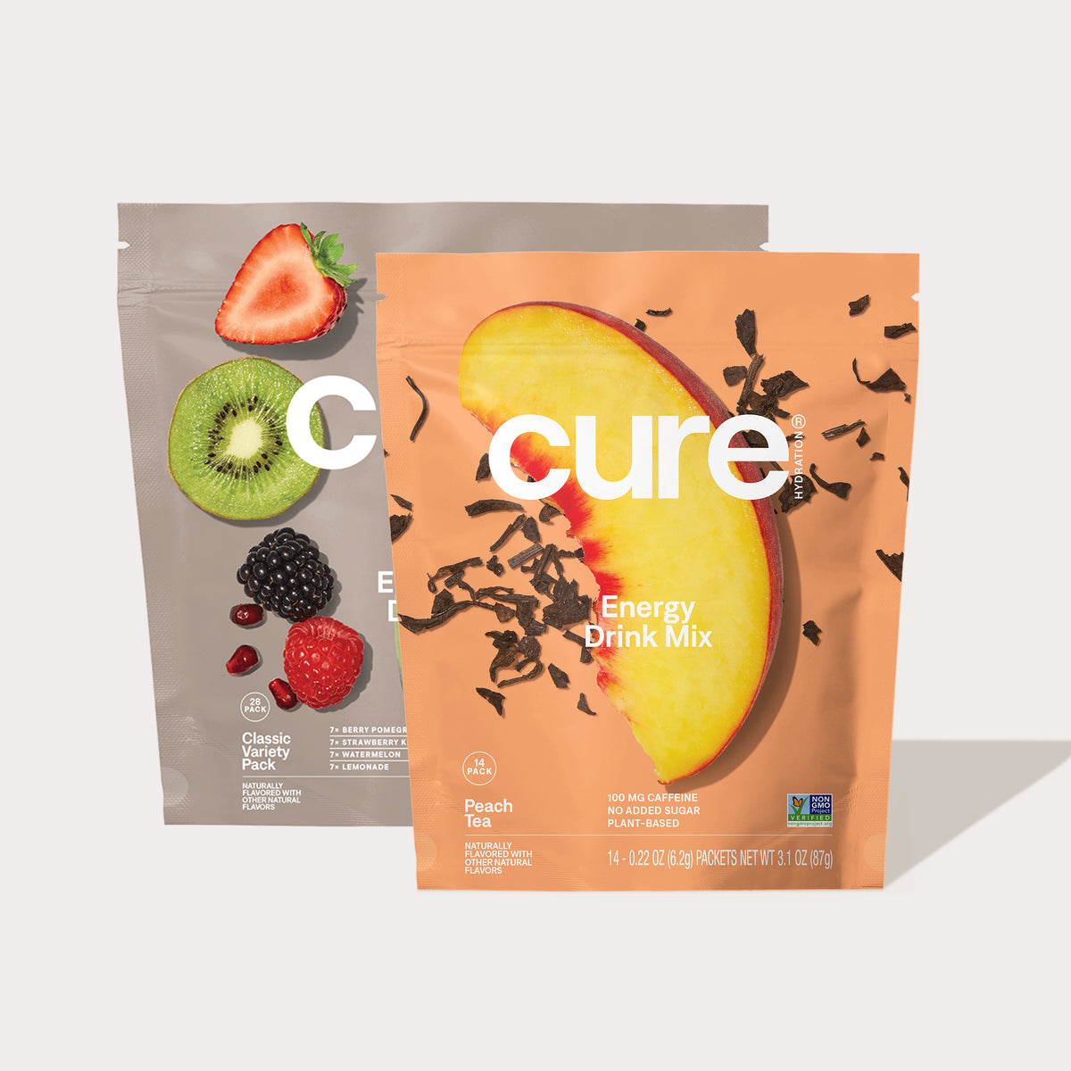 Two Cure Energy Drink Mix packages: one Classic Variety Pack with images of strawberry, kiwi, blackberry, raspberry, and pomegranate seeds, and one Peach Tea pack with a large peach slice and loose tea leaves.