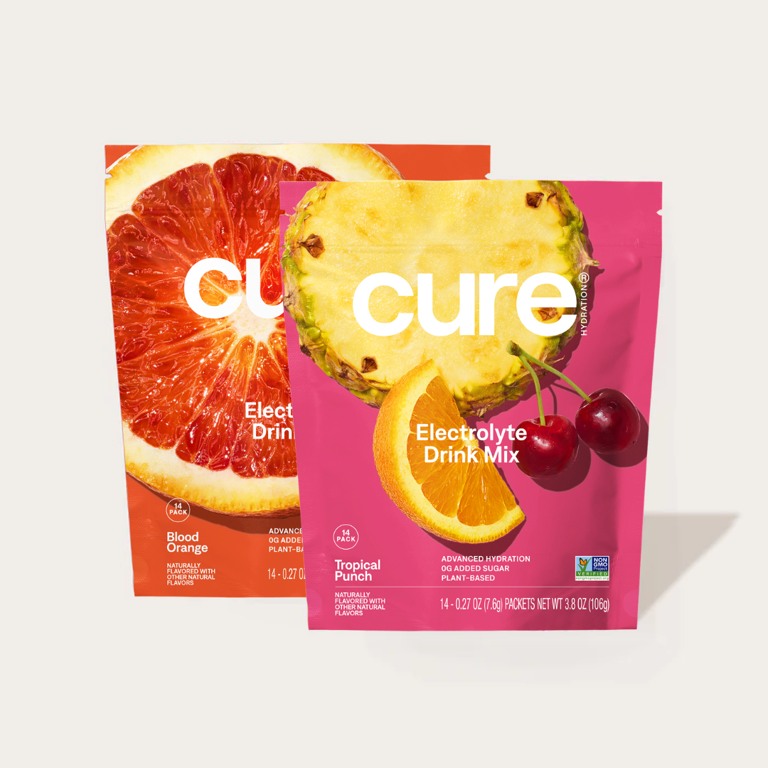 Two Cure Electrolyte Drink Mix packets, one Blood Orange flavor and one Tropical Punch flavor, with images of blood orange, pineapple, orange slice, and cherries on the packaging.