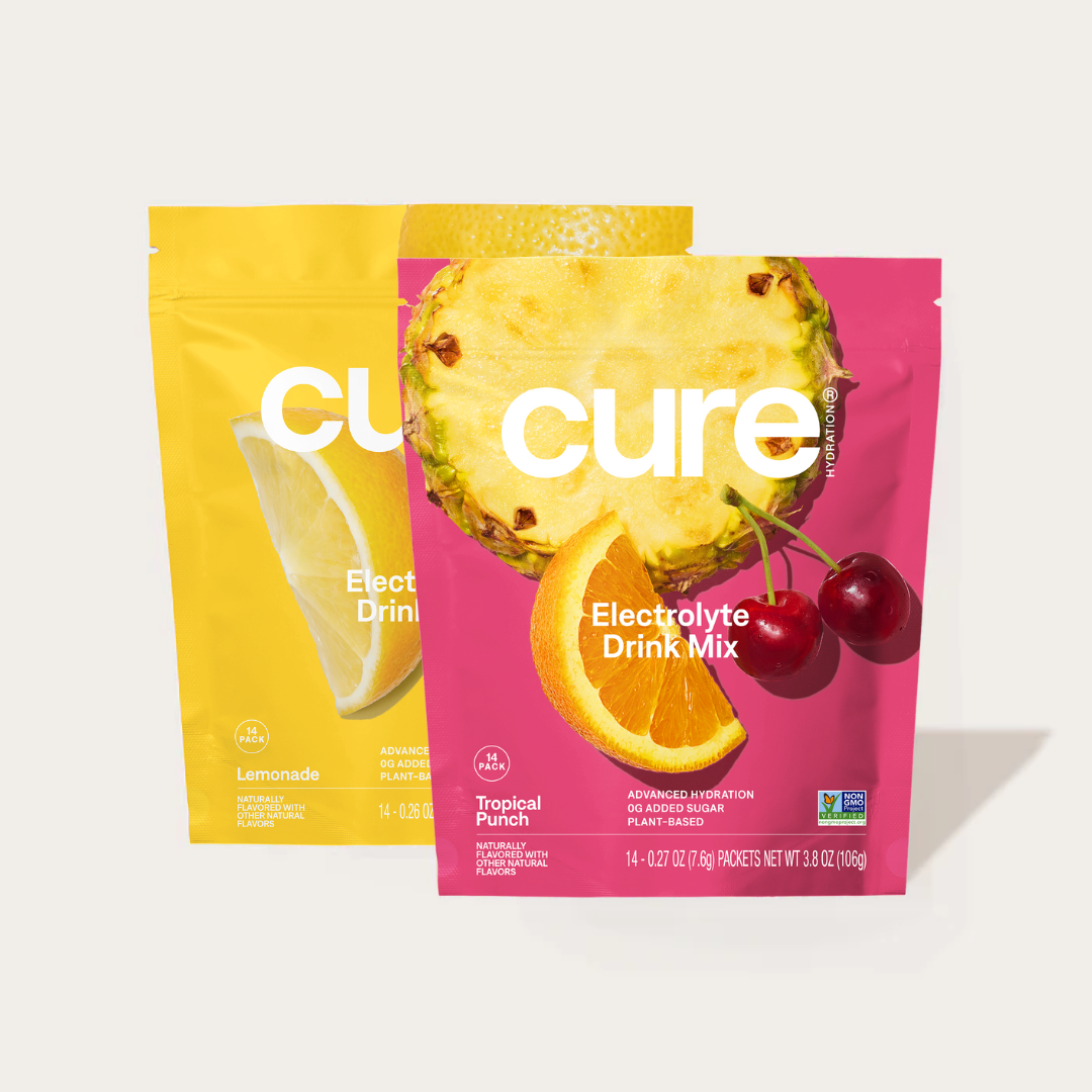Two electrolyte drink mix pouches labeled 'cure': one Tropical Punch flavor with pineapple, orange, and cherry images on pink packaging, and one Lemonade flavor on yellow packaging with a lemon slice image.