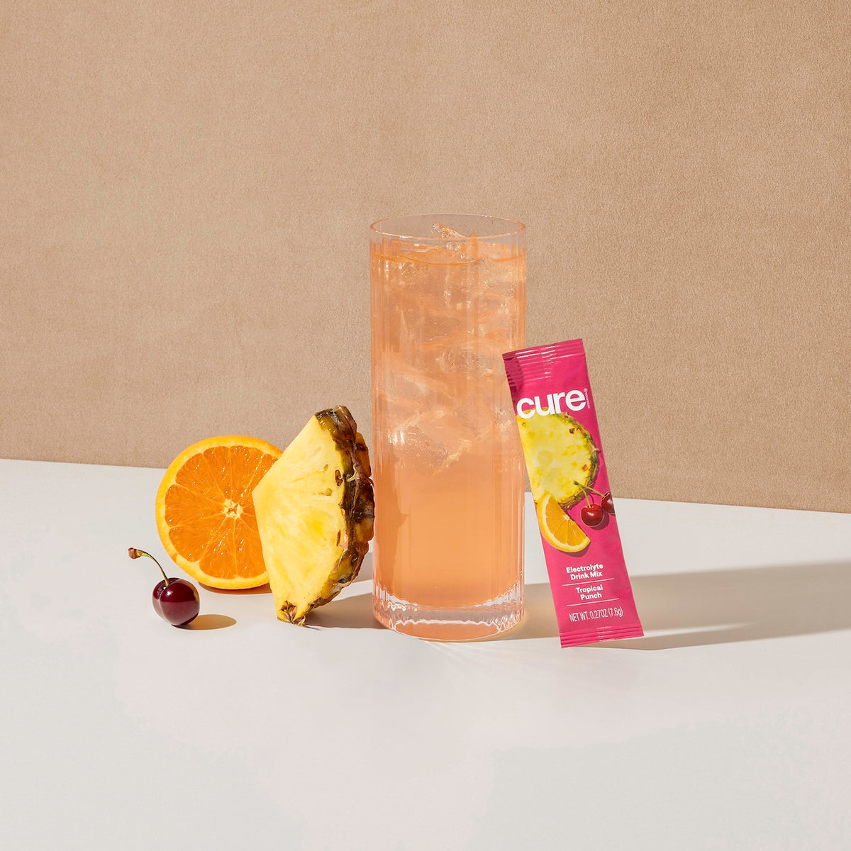 Tall glass of pink tropical punch drink with ice, next to a cherry, half an orange, a pineapple slice, and a pink Cure packet labeled 'Tropical Punch Berry Pomegranate'.