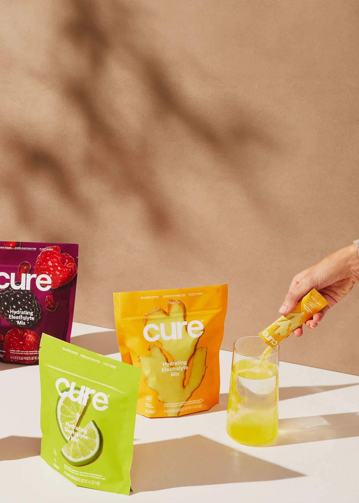 'Cure' electrolyte packets and drinks with a hand pouring a yellow mix, neutral backdrop