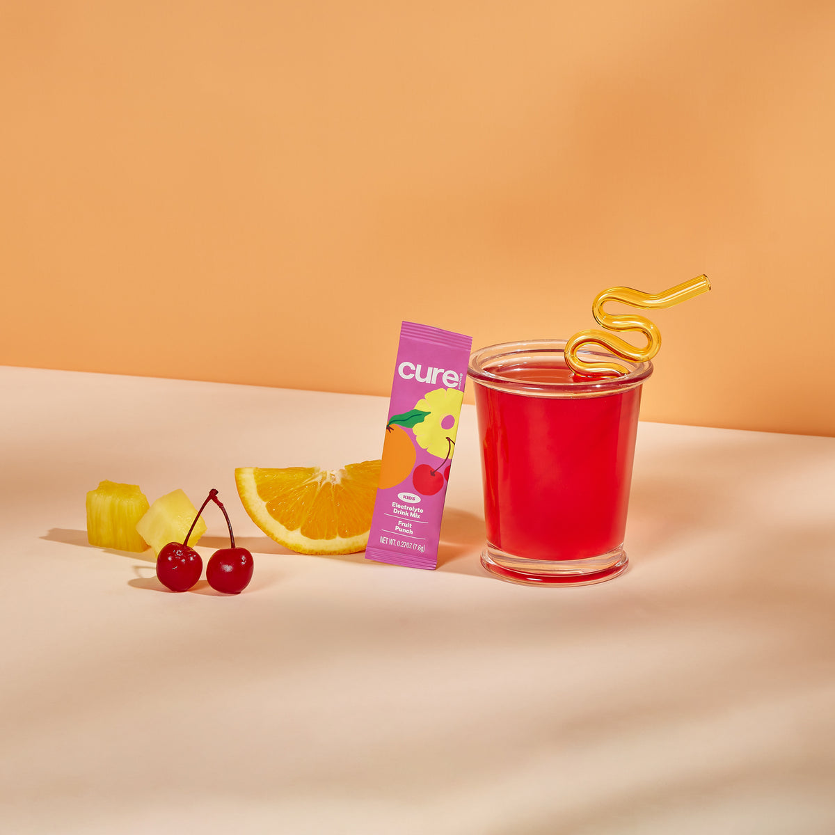 Red fruit punch drink in a glass with a yellow curly straw, Cure Fruit Punch product box, and fresh fruits including cherries, pineapple chunks, and an orange slice on a beige surface.