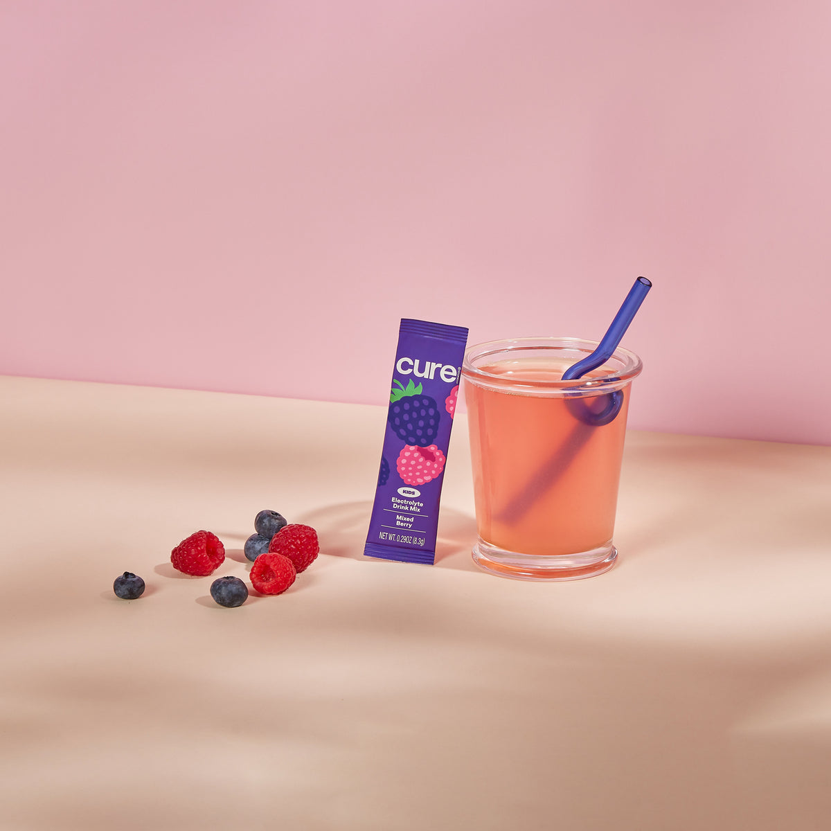 Glass of pink mixed berry drink with a blue straw, a purple box labeled 'cure' with a raspberry image, and scattered blueberries and raspberries on a beige surface.