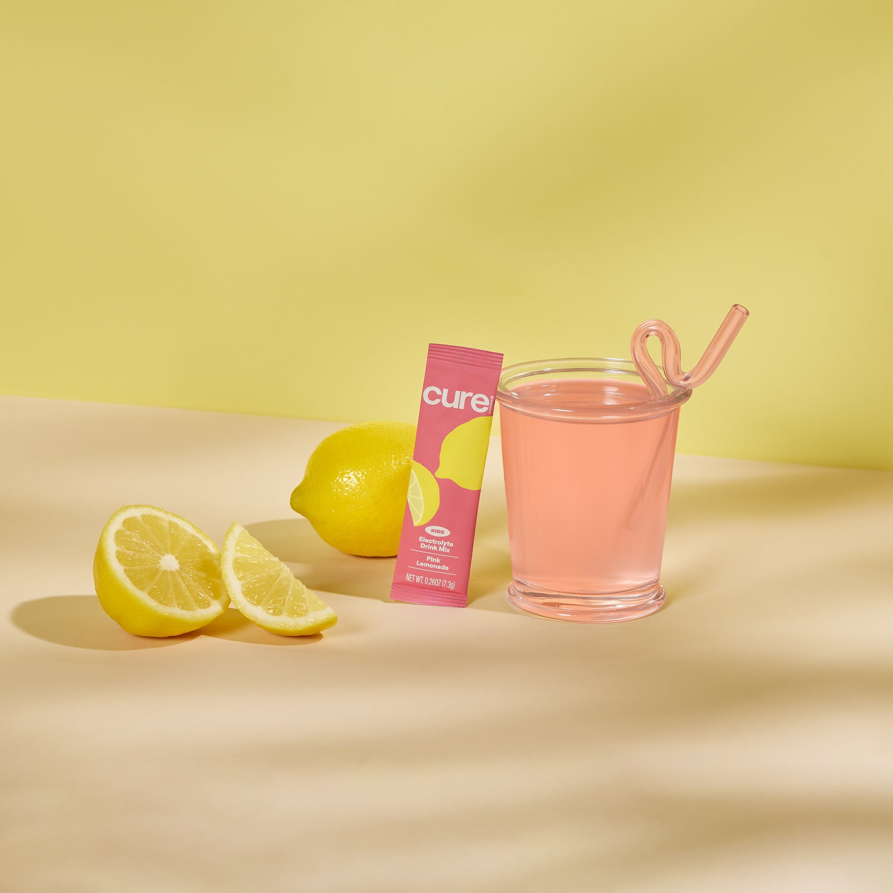 Glass of pink drink with a pink 'cure' packet and lemon slices on a yellow background