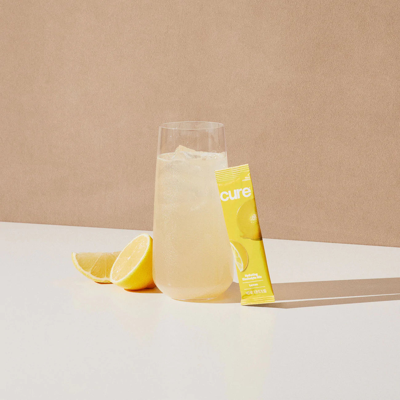Glass of lemonade with ice, lemon slices, and a yellow 'cure' packet on a white surface with a beige background