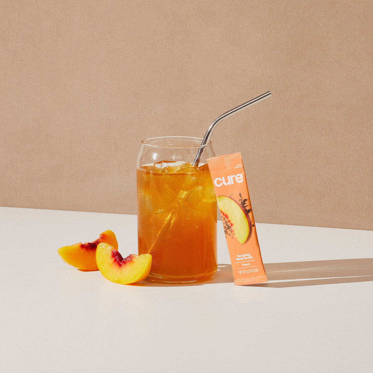 Glass of iced peach tea with metal straw, two peach slices, and a peach-flavored tea packet labeled 'cure'