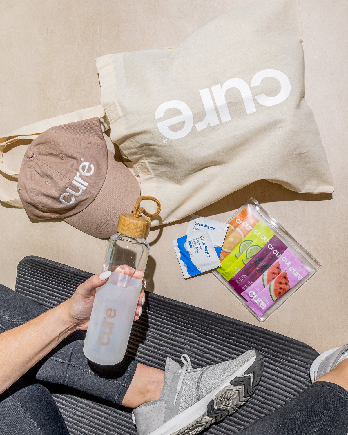 A beige Cure-branded hat, a tote bag with the word 'cure' printed in white, a clear water bottle with a wooden cap labeled 'cure', and wellness product packets laid out on a mat next to a person wearing gray sneakers and black leggings.