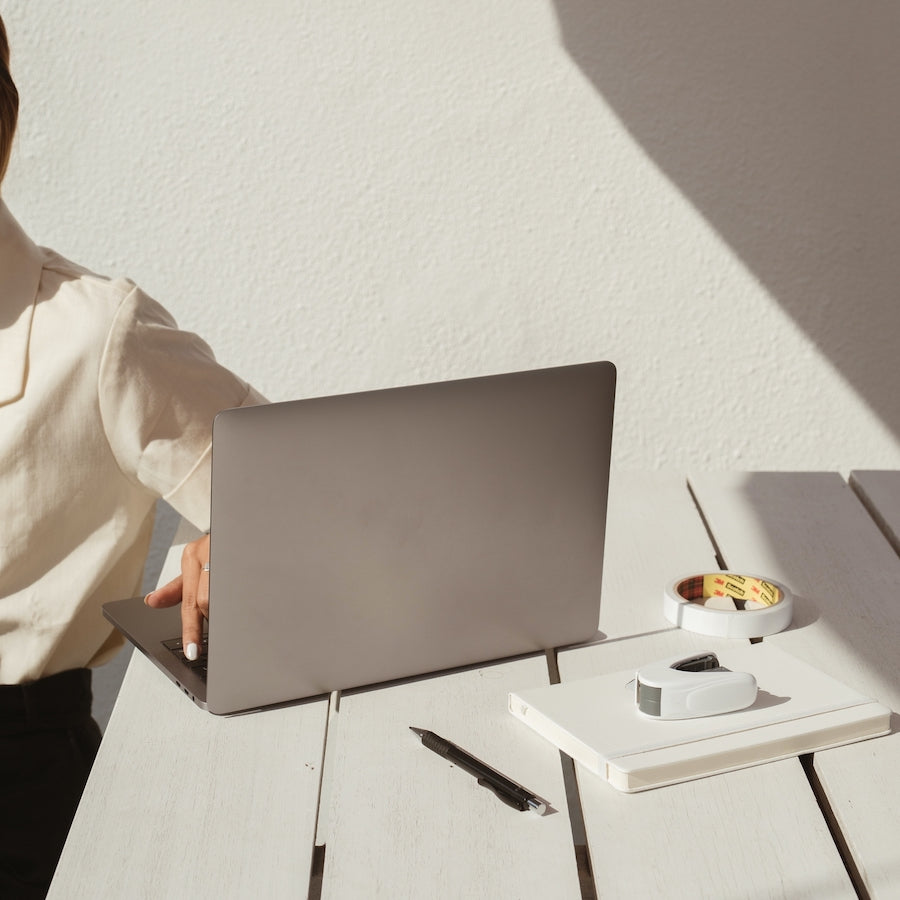 Person typing on a laptop at a sunlit white wooden table with a notebook, pen, and roll of tape