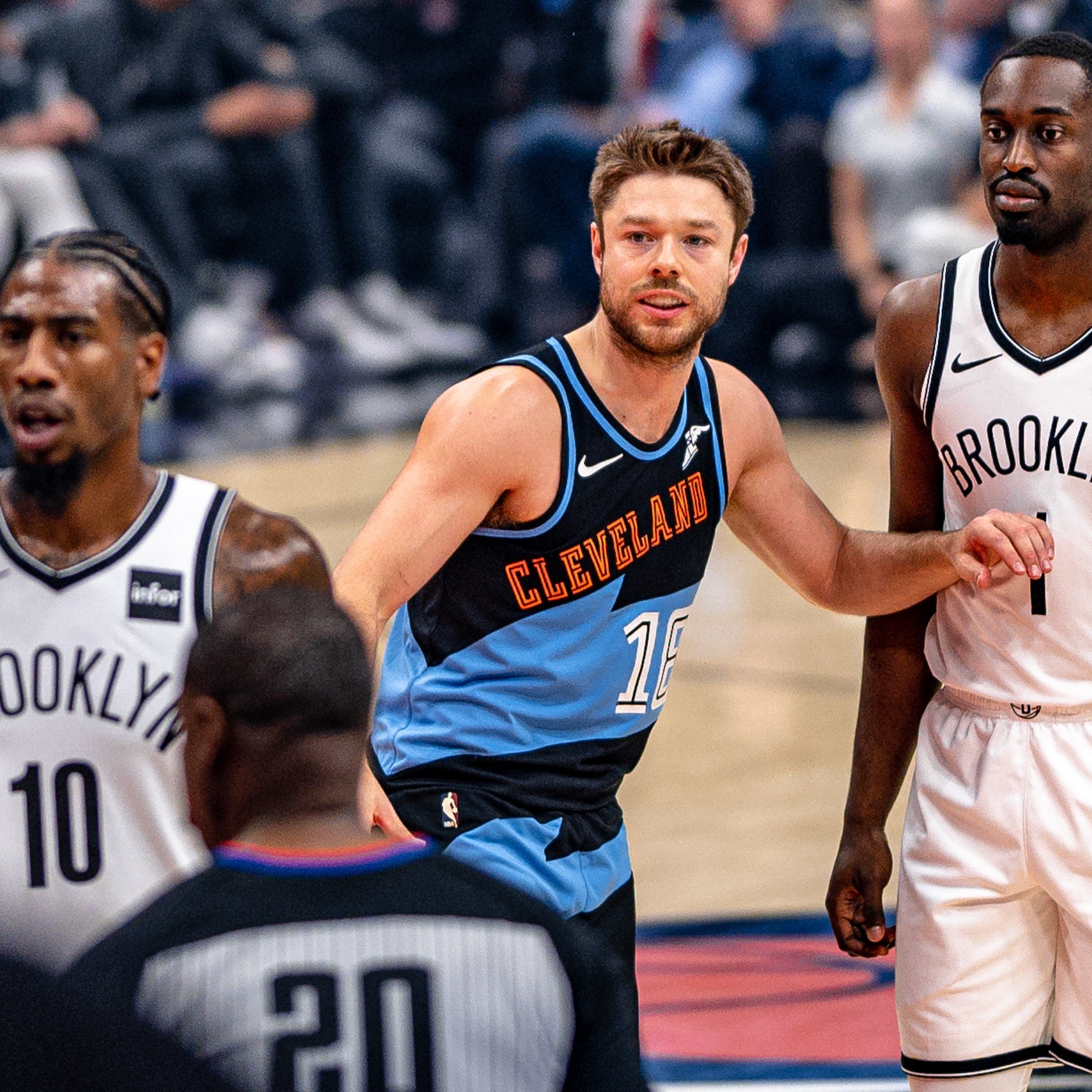Cleveland player defending between two Brooklyn Nets players on court with referee and blurred crowd in background