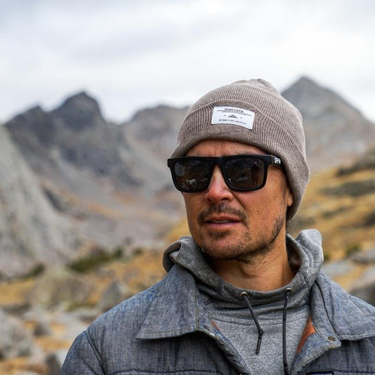Man wearing a gray beanie and sunglasses standing in a rocky mountain landscape