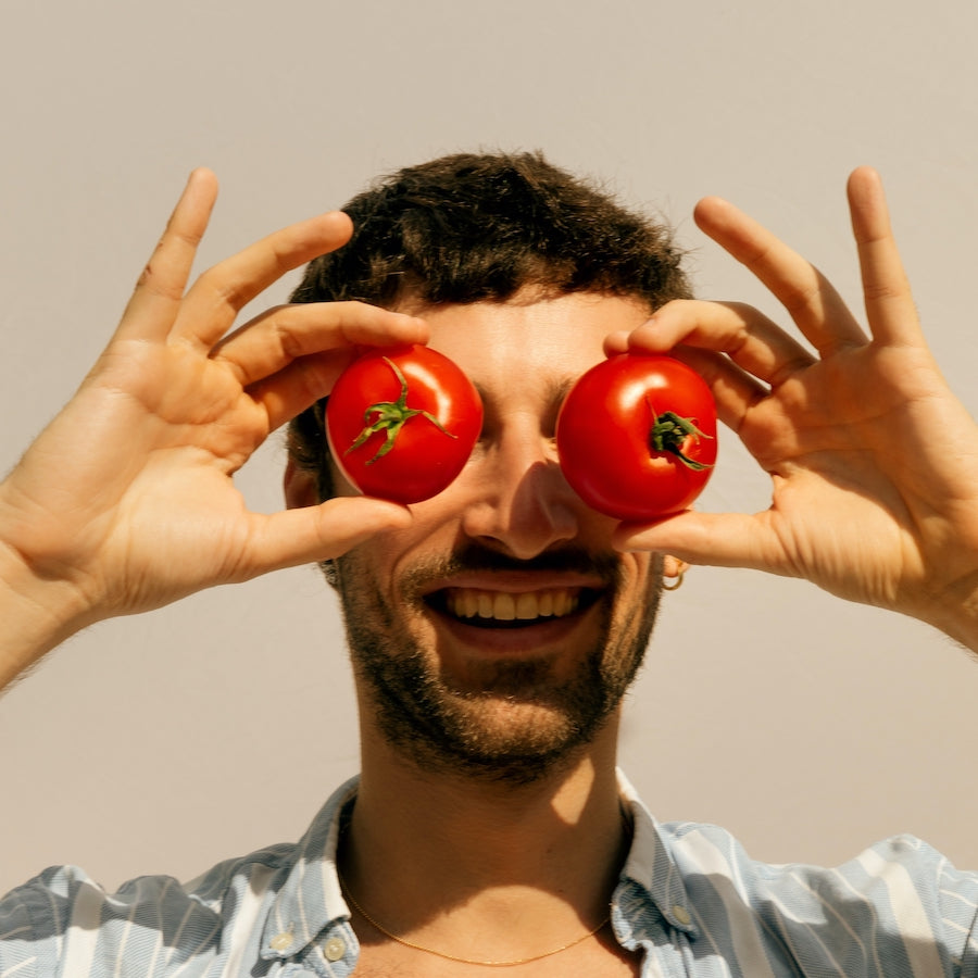 A smiling man holding two red tomatoes over his eyes against a plain background.