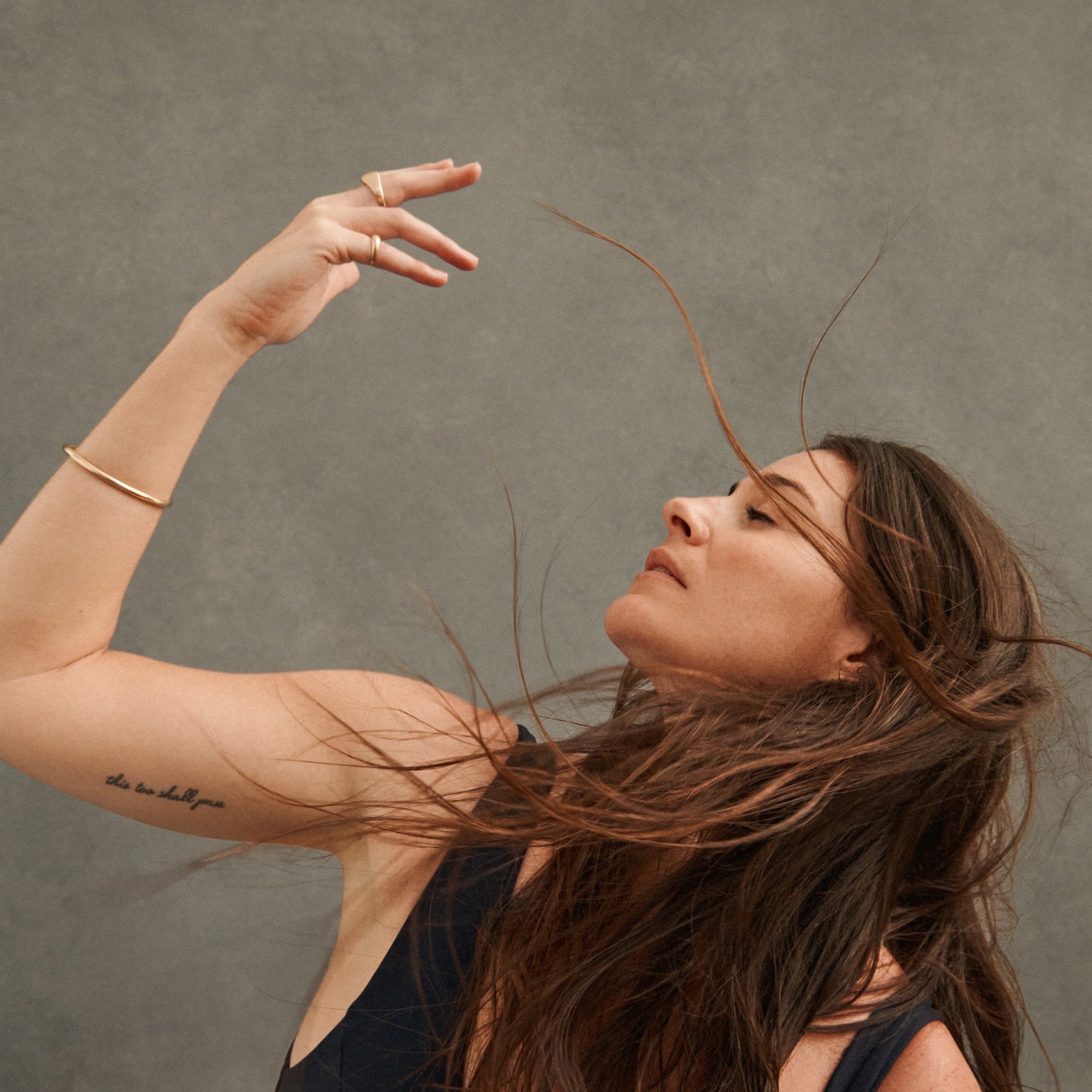 Raj Barker in profile, arm raised, hair swept back against gray background, inner-arm tattoo this too shall pass