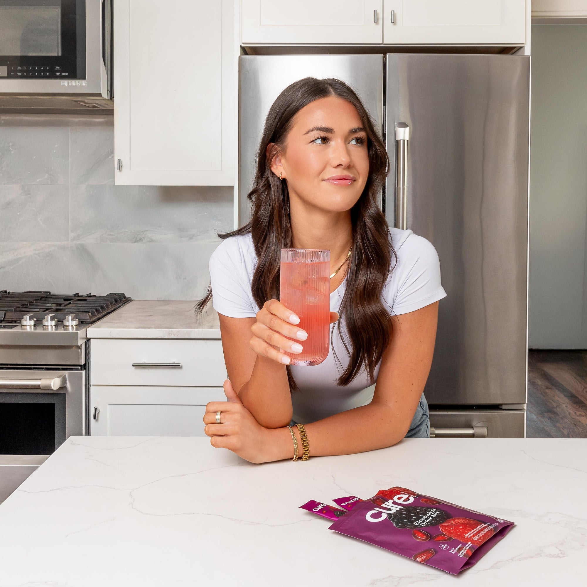 Woman holding a pink drink in a kitchen with Cure electrolyte drink mix packets on the counter