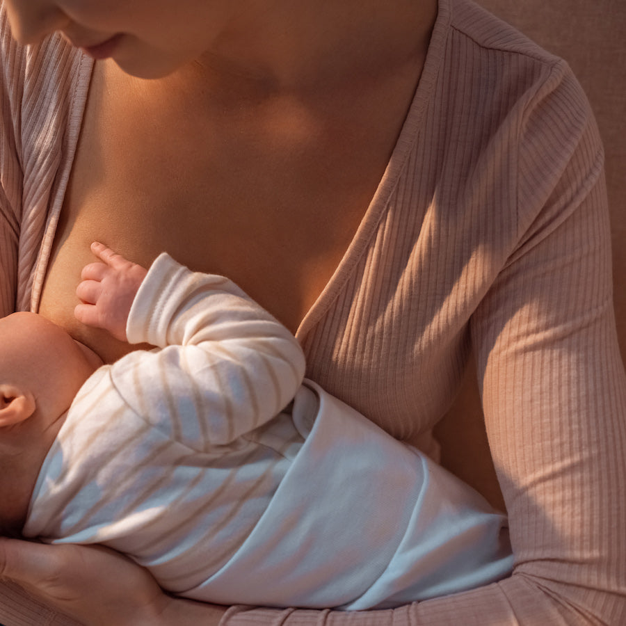 Mother breastfeeding newborn, close-up of baby latched to breast with mother in a ribbed pink top and warm indoor lighting