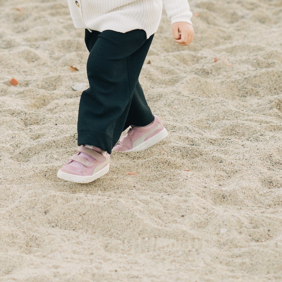 Toddler wearing black pants and pink sneakers walking on sandy ground