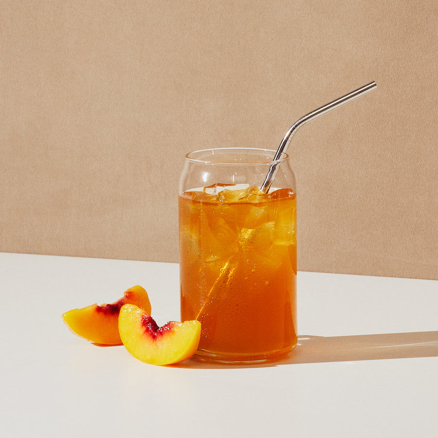 Glass of iced orange drink with metal straw next to two peach slices on white surface and beige background
