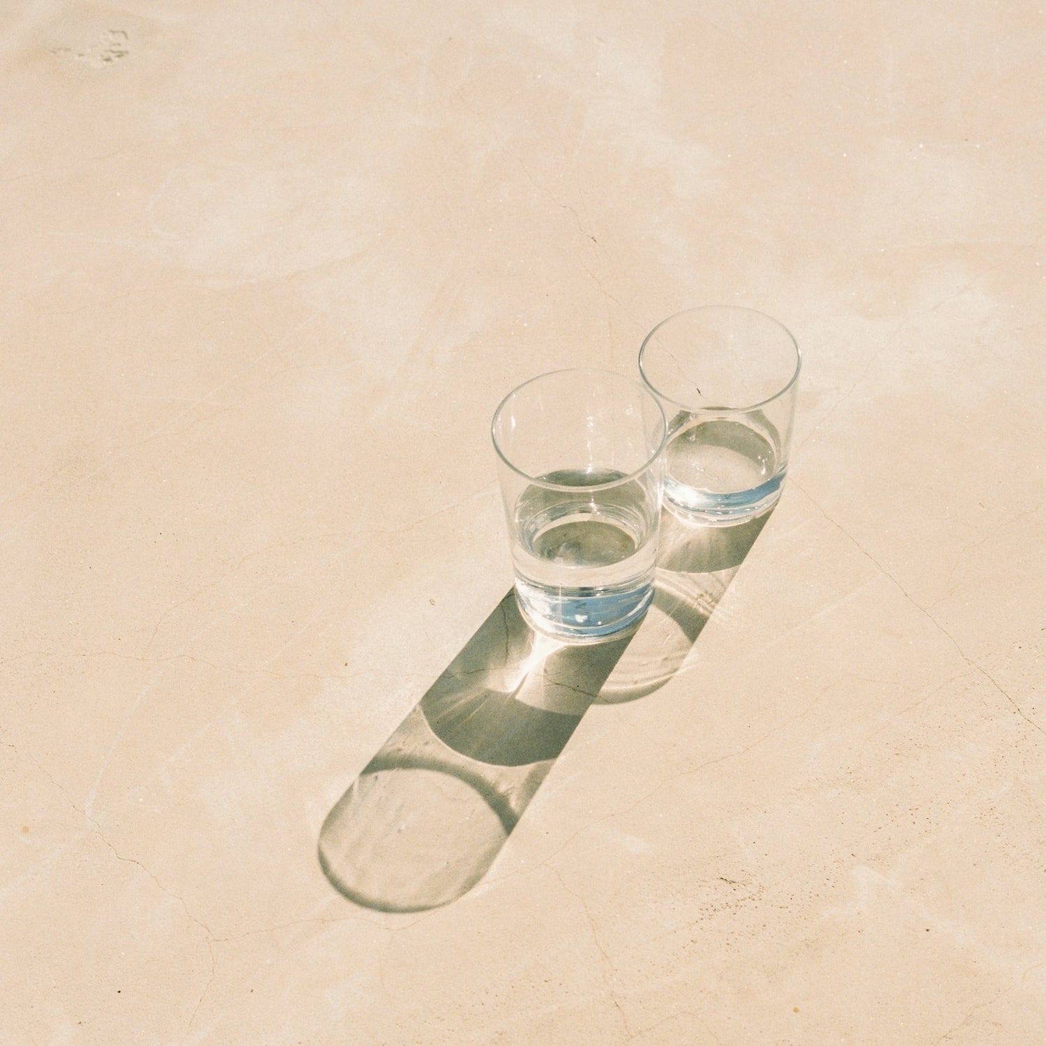Two clear glasses partially filled with water casting elongated shadows on a light beige surface under sunlight.
