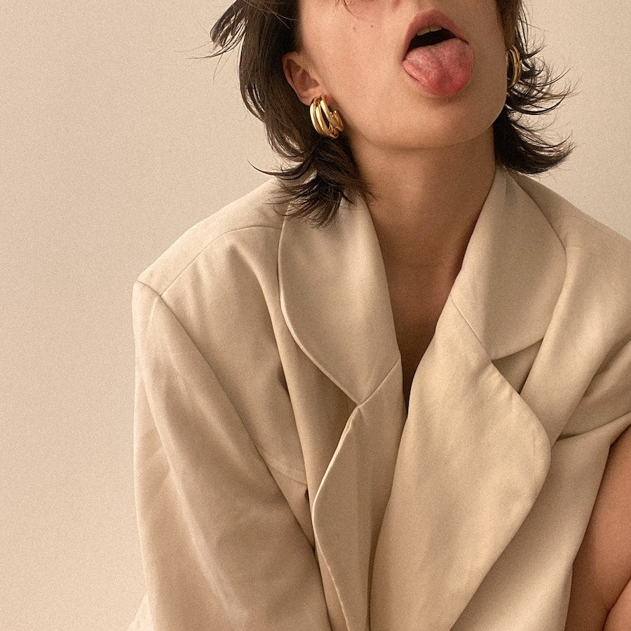 Woman in a beige blazer leaning forward and sticking out her tongue against a plain neutral background