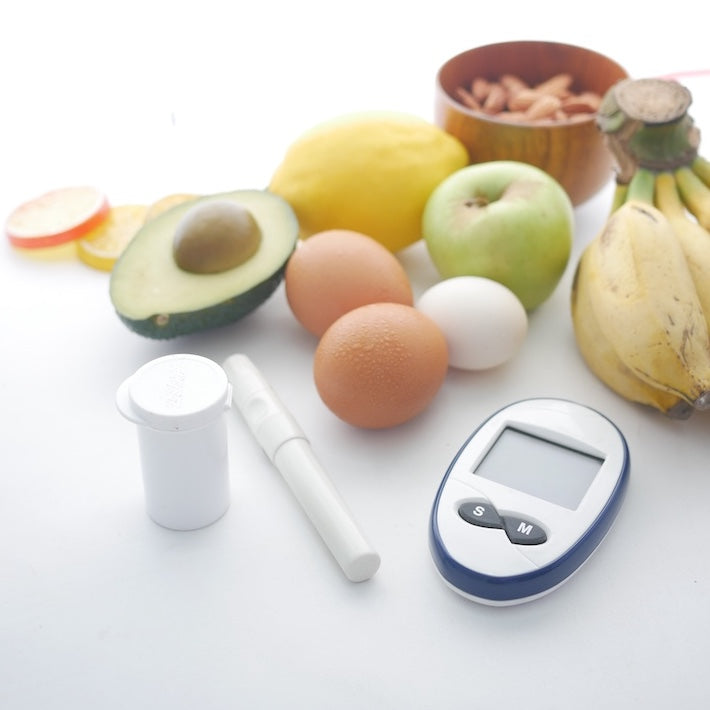 Glucometer, lancing device, and test strip container arranged with avocado, apple, eggs, bananas, and a bowl of nuts on a white surface