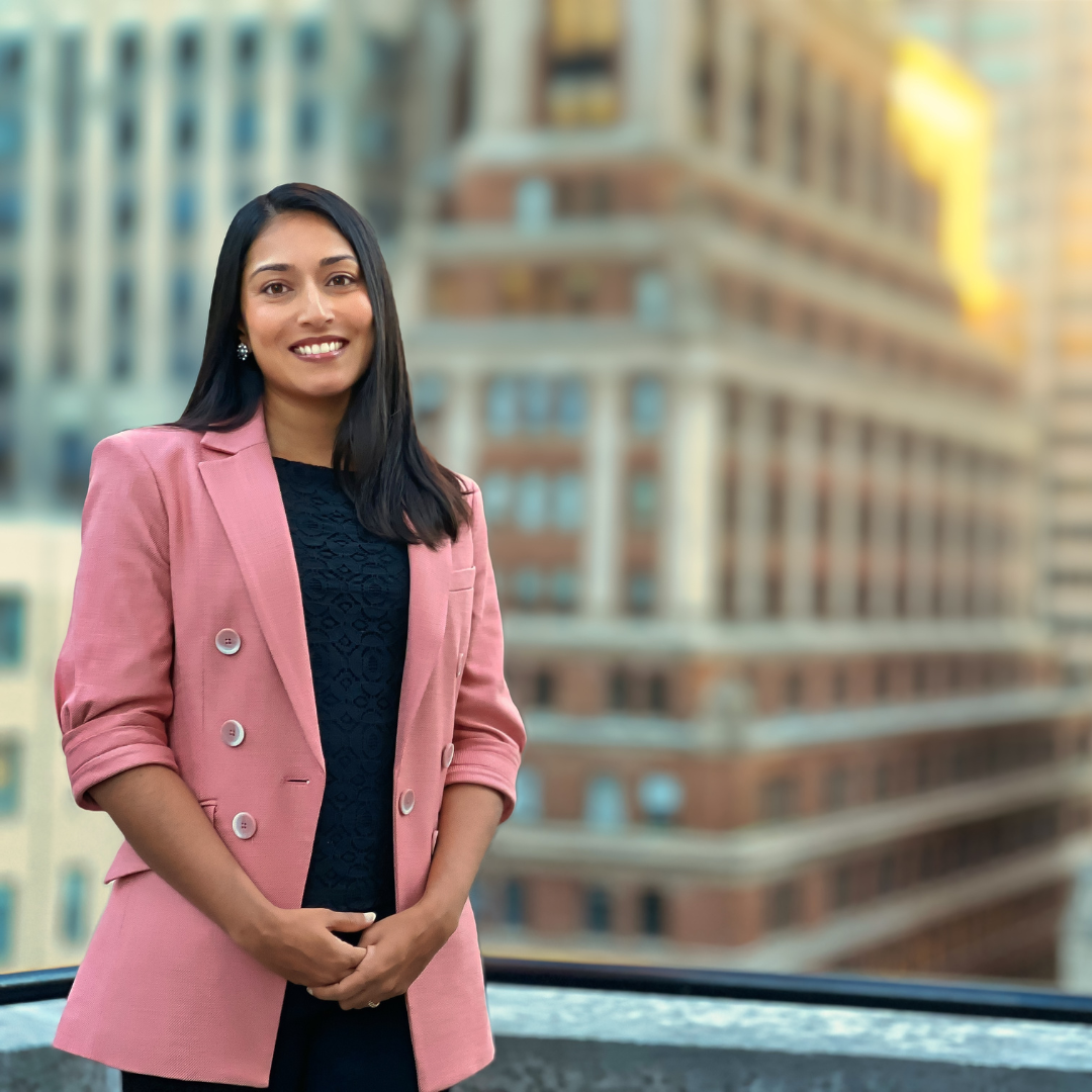 Dr. Arunima Agarwal smiling on a city rooftop wearing a pink blazer with blurred office buildings behind her