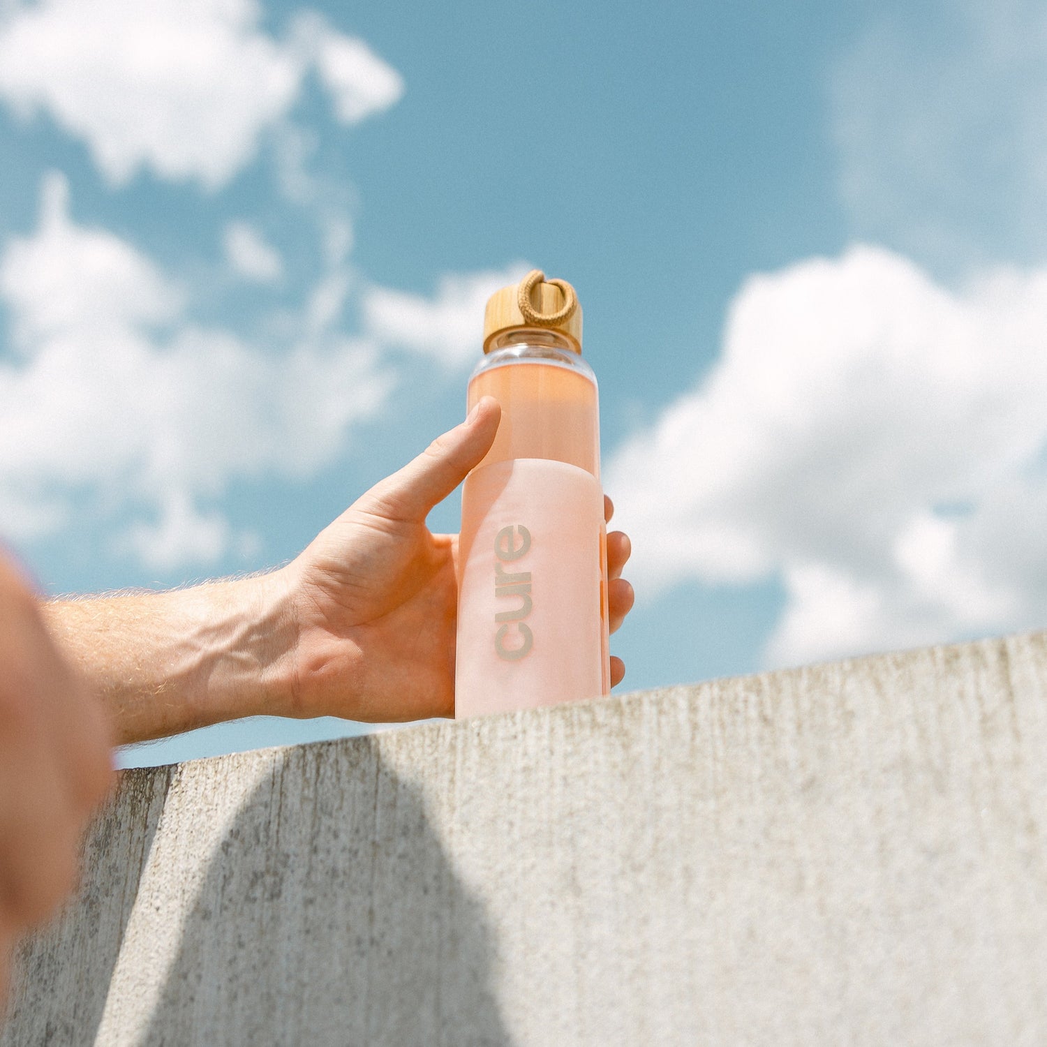 Hand holding a water bottle labeled 'cure' against a blue sky with clouds
