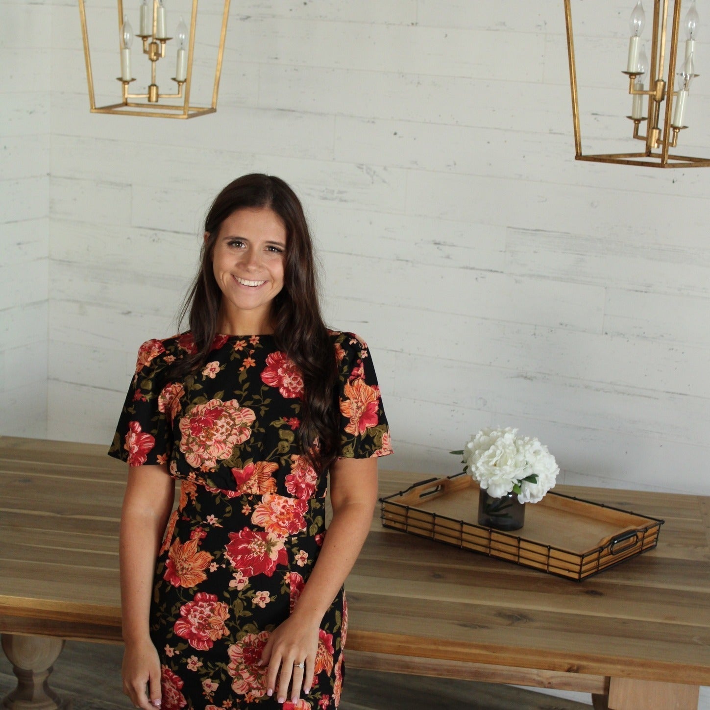 Smiling woman in a floral dress standing by a wooden table with a flower arrangement in a bright room 