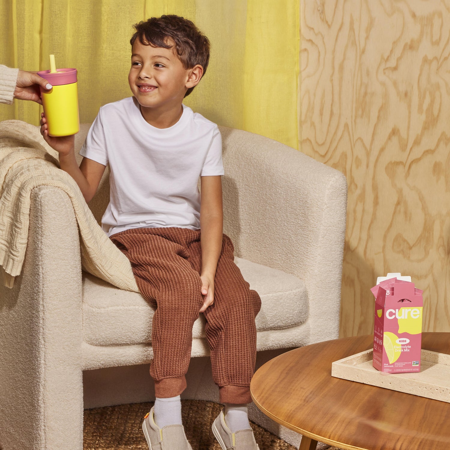 Smiling boy on an armchair taking a yellow cup; pink carton on table labeled cure, KIDS, Electrolyte Drink Mix.