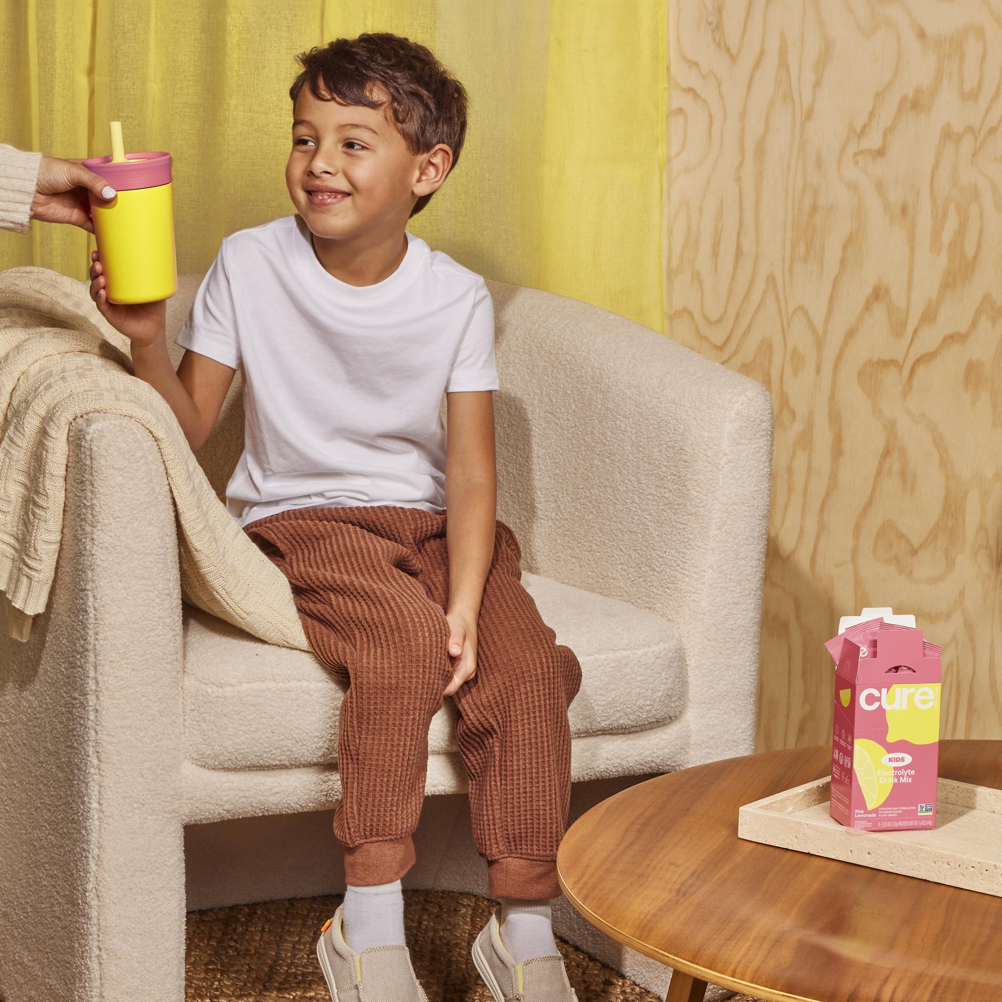 Smiling boy on an armchair taking a yellow cup; pink carton on table labeled cure, KIDS, Electrolyte Drink Mix.
