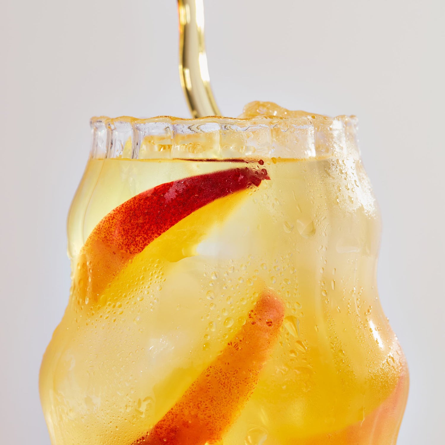Close-up of a glass of peach mango electrolyte drink being poured with ice and peach slices against a pale background